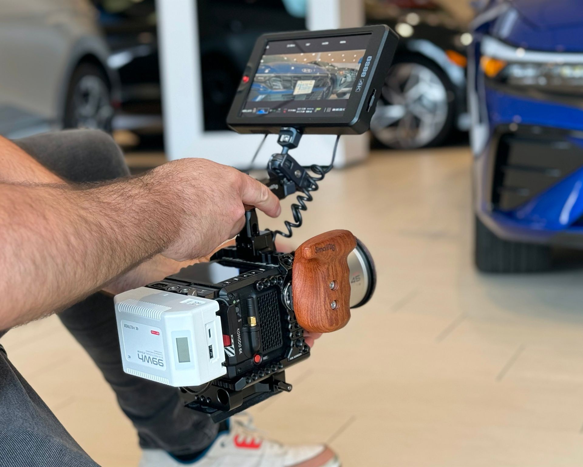 Person holding a professional video camera rig with a monitor, filming a blue car in a showroom.